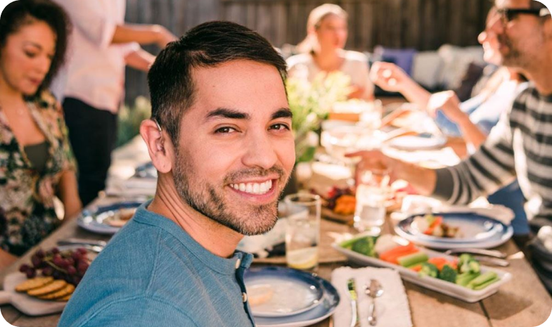 A man looks over his shoulder, smiling at the camera. He sits at a picnic table with friends, sharing a meal.