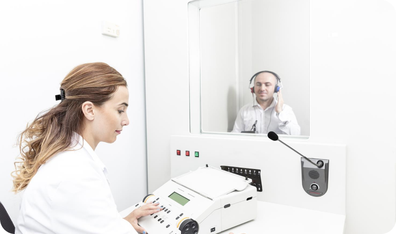 A woman in a white coat uses a machine to test the hearing of a man wearing headphones in a testing booth. 