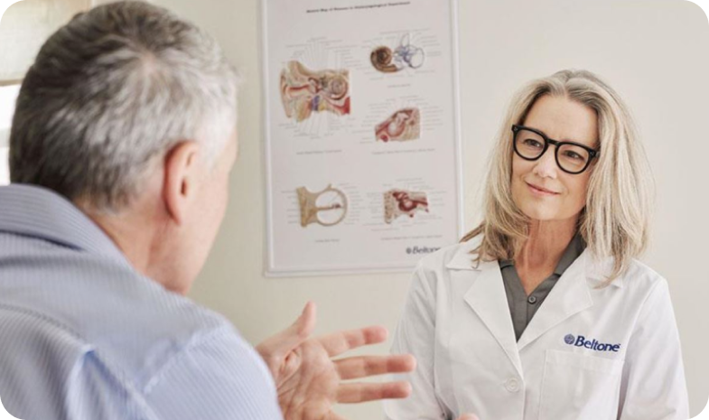 An older man in a light blue shirt uses hand gestures to speak with a Beltone practitioner wearing a white jacket and glasses. 