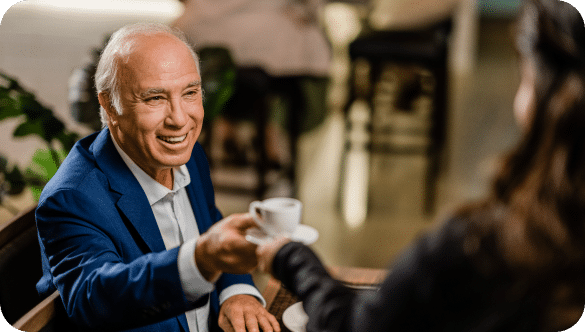 An older man accepts a cup of tea from a woman, while exchanging a smile.