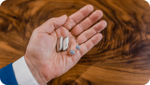 A person hold a pair of Betone Achieve hearing aids in the palm of their hand. The hearing aids are small gray receivers with short cords attached to small gray ear buds.