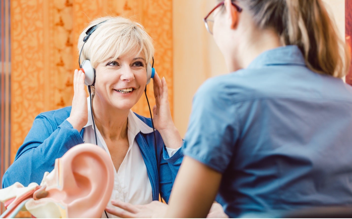 Two people in blue sit across from each other for a hearing test; one person is wearing headphones, and there is a model of an ear in the foreground.