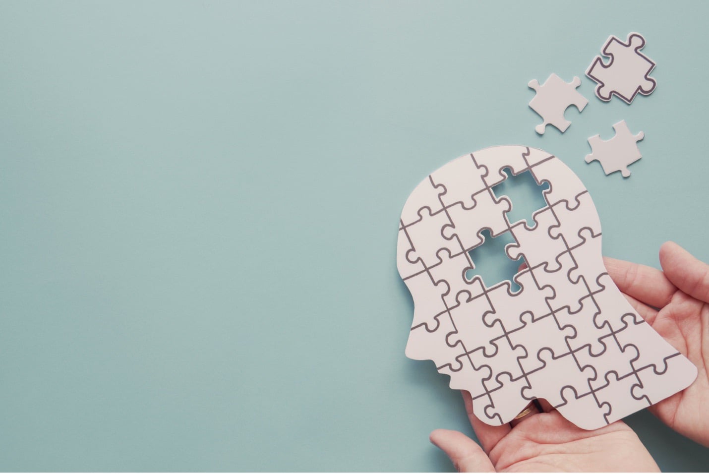 A person’s hands hold a puzzle shaped like a human head; the white puzzle has two pieces missing from the center, and the image is on a light blue background.