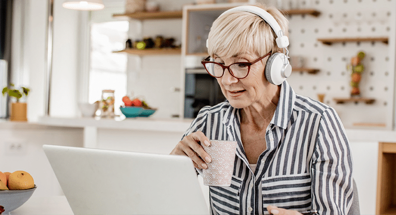 An older woman sits at a laptop, wearing headphones and drinking from a cup of coffee. She is taking a free online hearing test.