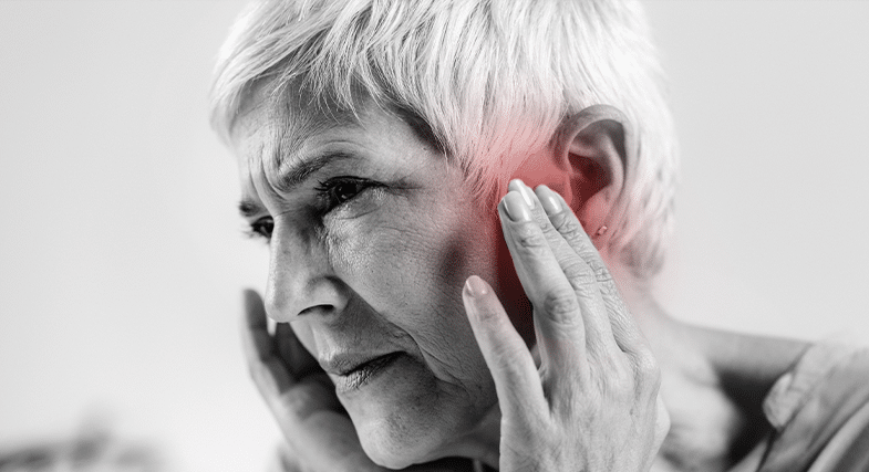 A black and white image of an older woman with short, straight white hair shows her pressing her fingertips to the front of her ears, where the photo is tinted red. 