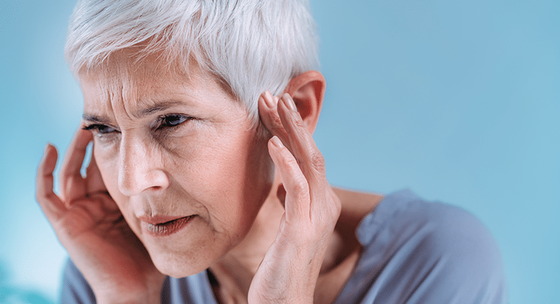 An older woman with short white hair holds her hands up to her ears, pressing on the front of her ears with her fingertips. 