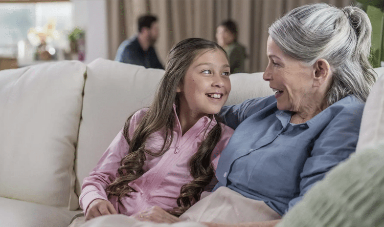 An older woman and a younger girl sit side-by-side on a couch, chatting and smiling at each other.